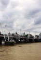 A view of the Thames river looking very brown under a clouded steel-grey sky. the Golden Jubilee Footbridges underline the background disney-style buildings