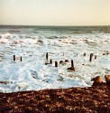 a golden pebble beach in the foreground has evening shadows, but the foaming sea is still catching the sun and the background sky is still light. Small rocks and short, weathered wooden posts protrude from the shallow sea foam.