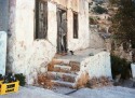 crumbling steps up to a dilapidated building where a black and white cat stands at the rotted wooden door. In the left corner of the picture a yellow plastic crate overflows with empty bottles.